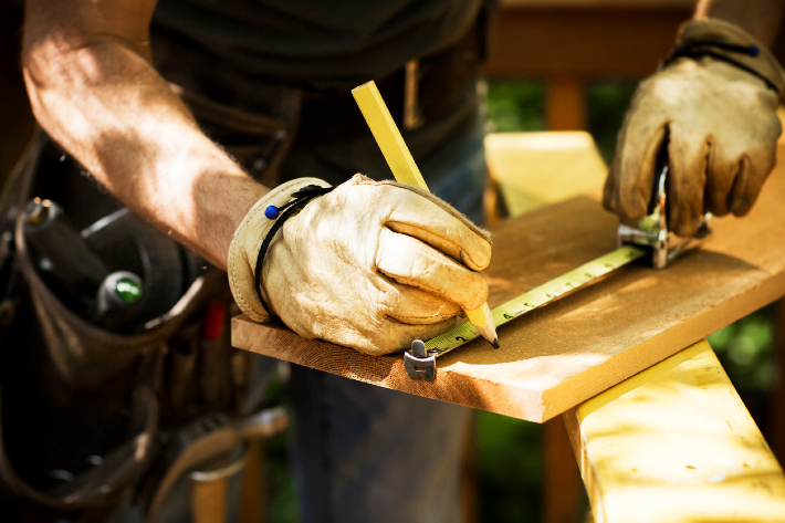 A man wearing woodworking gloves while measuring a plank.